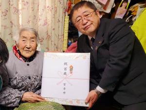 椅子に腰かけた100歳を迎えた吉田たかさんと、中腰になった市長が「祝 百歳」と書かれた熨斗がかかった贈呈品を一緒に持ち写っている写真