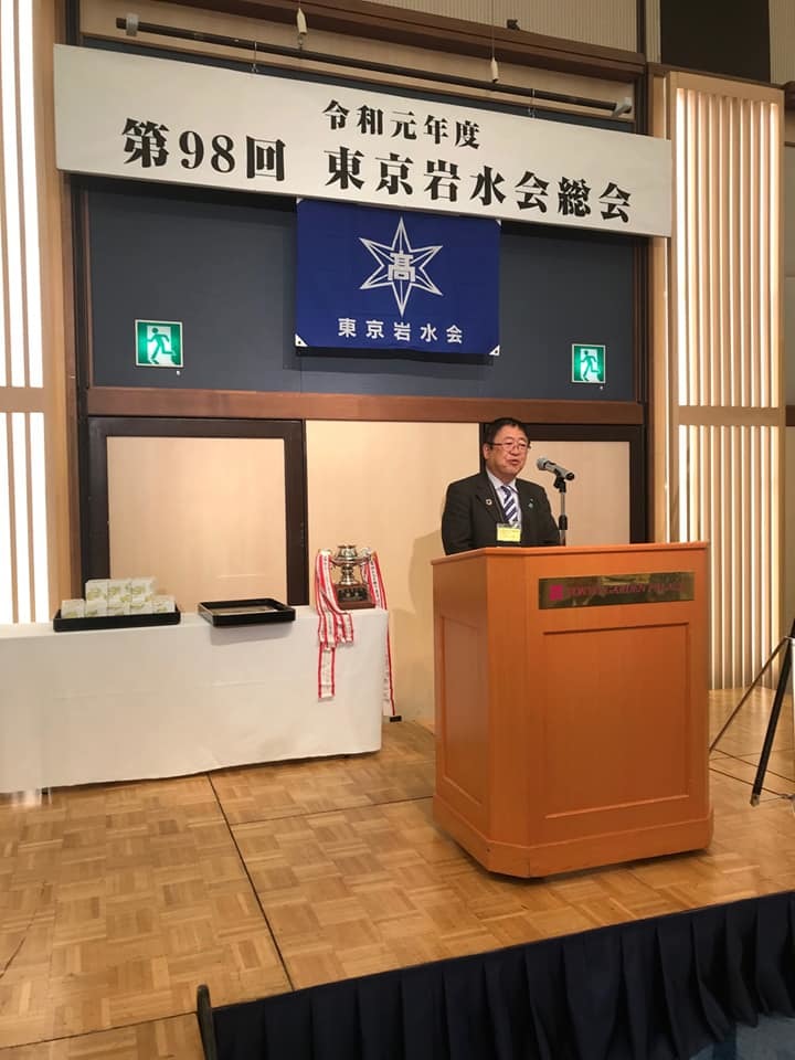 「令和元年度第98回東京岩水会総会」と書かれた横断幕が吊るされた舞台上で、演台の前で話をする様子の市長