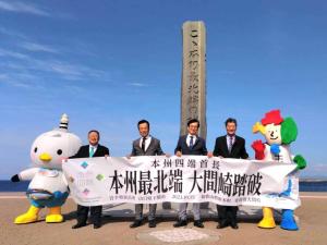 海岸沿いに立つ、「ここ本州最北端の地」の碑の前に4人の首長とゆるキャラの「かもまーる」「よんたん」が「本州四端首長 本州最北端 大間崎踏破」と書かれた横断幕を持って並んでいる記念写真