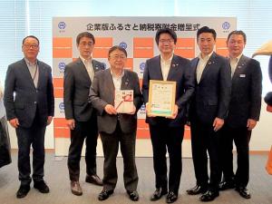「企業版ふるさと納税寄附金贈呈式」と書かれたバックパネルの前で、目録を持った市長と額縁を持った男性を中心に、両側に2名ずつ関係者の男性が立ち横一列に並んでいる写真