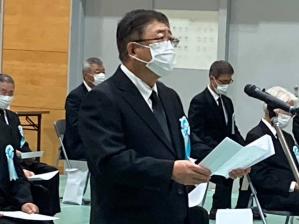 多くの参加者が座っている中、喪服のスーツ姿の市長がマイクの前で、式辞用紙を読んでいる写真