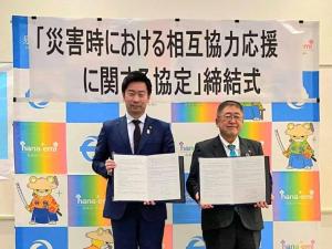 「災害時における相互協力応援に関する協定締結式」と書かれたバックパネルの前で、市長と男性がそれぞれ協定書を手に持っている写真