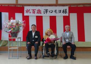 紅白幕の上に「祝百歳 沢口モヨ様」と書かれた横断幕が掲げられ、その前方のスペースで車いすに乗った満100歳を迎える澤口モヨさんを中心に市長と関係者の男性が椅子に座っている記念写真