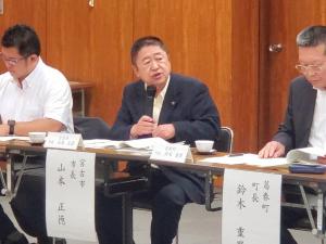 長机に名前が書かれた紙が貼られた席に市長が座り、両脇の男性が資料を見ている中、マイクで話をしている写真