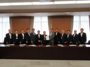要望書を一緒に持つ市長と男性1名、関係者の男性11名が窓際に横一列に並んでいる記念写真