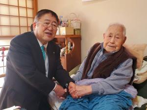 自宅の部屋で座っている満100歳を迎える影田久保伊勢次郎さんと両手で握手をしている市長の写真