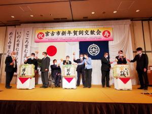 「宮古市新年賀詞交歓会」と書かれた横断幕が吊るされた壇上で、3つ置かれた酒樽を鏡開きする様子の参加者たちの写真