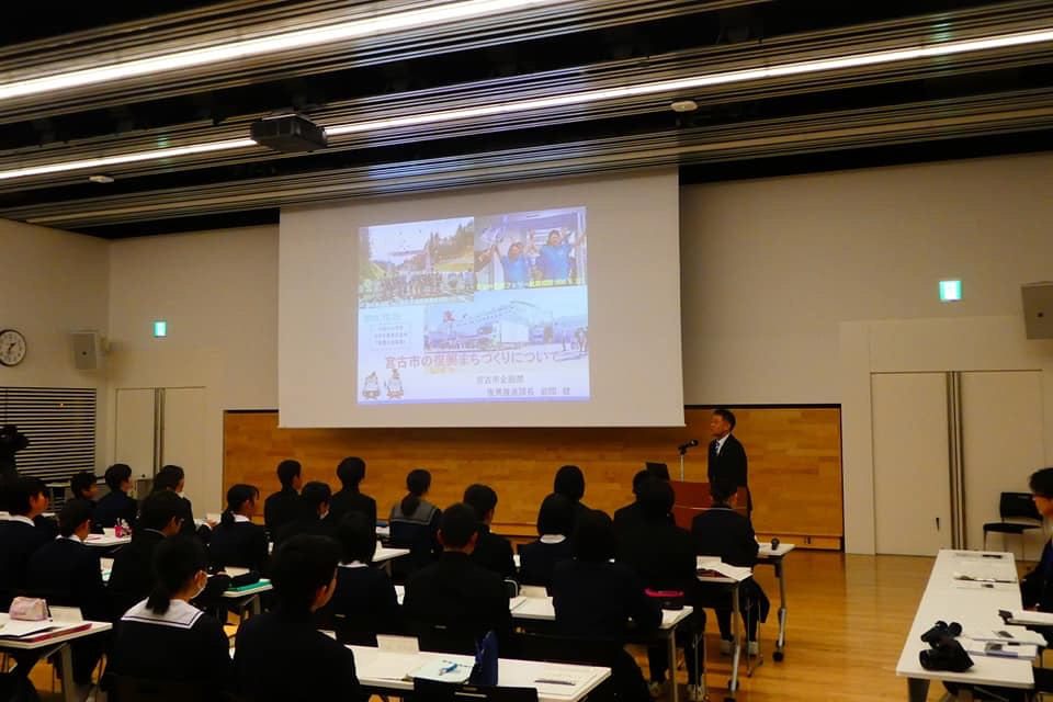 画像が投影されたスクリーンを見る様子の学生たちと、スクリーンの横に置かれた演台の前で話をする様子の男性の写真