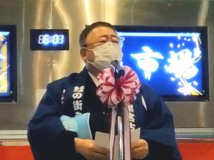 電子時計とモニターが設置された施設で、法被を着た市長が紅白リボンが付けられたスタンドマイクの前で話をする様子の写真