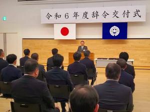 「令和6年度辞令交付式」と書かれた横断幕、国旗、市旗が掲揚された会場で、市長が座っている職員の方々の前で話をされている写真