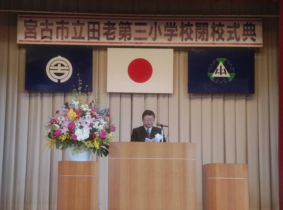 「宮古市立田老第三小学校閉校式典」と書かれた横断幕と宮古市の市町村旗、日本国旗、校旗が掲げられた舞台で、演台に置かれたマイクに向かい話をする様子の市長