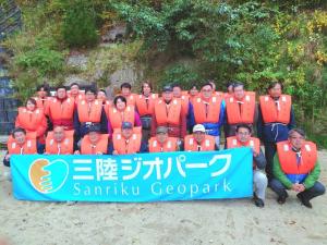 水色地の「三陸ジオパーク」と書かれた横断幕を持ち、ライフジャケットを着用した参加者が3列に並んでいる集合写真