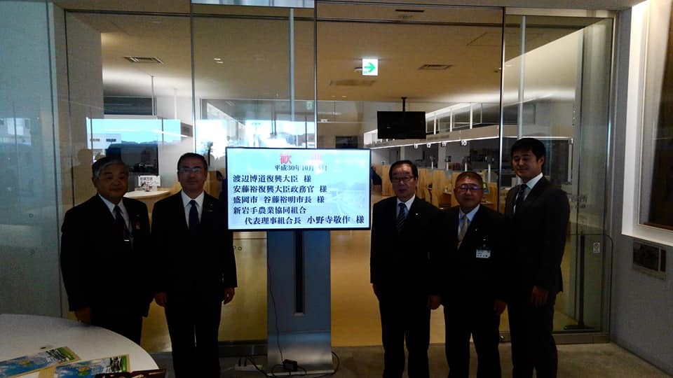 「歓迎」という文字と訪問される方たちの名前が映し出されたモニターの両側に立つ市長や渡辺博道復興大臣、安藤裕復興大臣政務官(兼内閣府大臣政務官)、関係者の方々計5名を写した写真