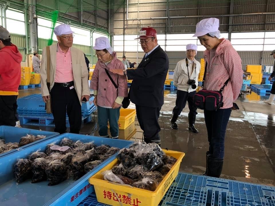 水色や黄色のバットに入った水揚げされた魚介類の傍に立つ観光客に、赤色のキャップを被った市長が説明を行っている写真