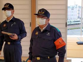 防災時の帽子を被り、作業着を着た市長が黒色で本部と書かれたオレンジ色の腕章をして、 訓練に参加している写真