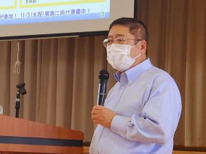 スクリーンが吊るされた会場で、右手にマイクを持ち話をしている様子の市長の写真