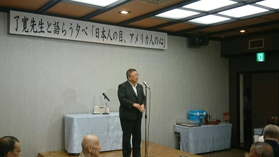 「「了寛先生と語らう夕べ「日本人の目、アメリカ人の心」」と書かれた横断幕が壁の上部に吊るされた会場で、スタンドマイクに向かい話をする様子の市長