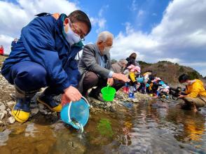 市長と他の参加者の方々が川沿いに並んで、バケツに入った稚魚を放流している写真