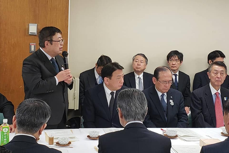 白色のテーブルクロスが掛けられたテーブルの席や壁側に並べた椅子に大勢の参加者が座っている会場で、自席から立ち上がった市長が両手でマイクを持ち話をしている様子の写真