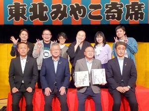 「東北みやこ寄席」と書かれた横断幕の下で、赤いシートが引かれた椅子の上に座る市長と3名の男性、その後ろで右手を広げたりピースのポーズをする演者の方達6名が写っている記念写真