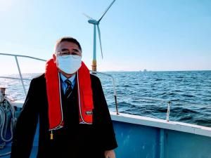 オレンジ色のライフジャケットを首にかけた市長を浮体式洋上風力発電施設を背景に船上で撮影した写真