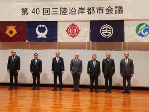 「第40回三陸沿岸都市会議」と書かれた幕の下で関係者7名が1列に並び記念撮影をしている写真