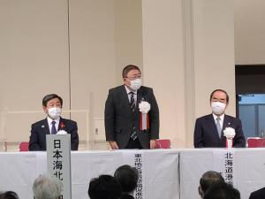 白いクロスが敷かれた机が置かれた会場で、机の後ろにつ白い胸章を着けて立つ市長と、市長の両側に座る白い胸章を着けた関係者の男性2名を写した写真