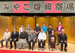 「みやこ復興寄席」と書かれた横断幕が掛けられ、金屏風が後方に立てられた舞台上で、椅子に座る市長や参加者の方々7名が前列、後列に6名の参加者の方々が立って並んでいる記念写真