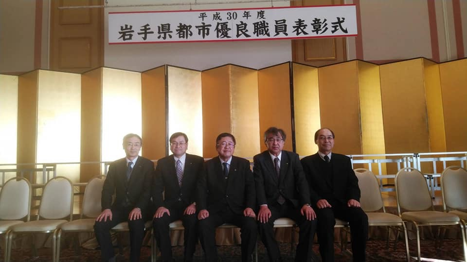 「平成30年度岩手県都市優良職員表彰式」と書かれた横断幕や金屏風が設置された会場に並んだ白色の椅子に、市長を中央に左右に2名ずつ4名の受賞者が横一列に並んで座っている写真