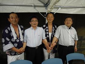 テントの下で、法被を着た小木田喜美雄秋田市議会議長と穂積志秋田市長と、石田達郎秋田市歯科医師会会長と市長が4人で交差して手を交わしている写真