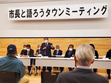 「市長と語ろうタウンミーティング」と書かれた横断幕の下、2列並んだ長机に座るスーツ姿の男性たちの中央で市長が立って話している様子を撮影した写真