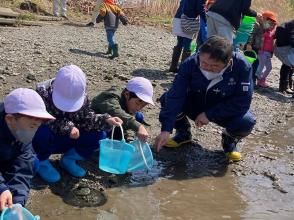 市長が3人の紫色の帽子を被った園児たちと川沿いで稚魚を放流している写真