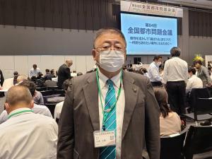「第84回全国都市問題会議」と書かれたスクリーンが前方にある会場で、椅子に座る参加者たちに背を向けて写っている市長の写真