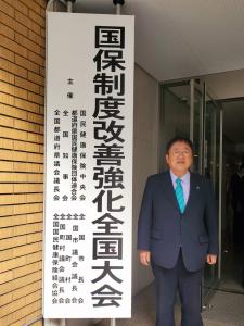 会場建物の入り口に設置された「国保制度改善強化全国大会」と書かれた看板の隣に立つ市長の写真