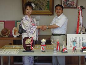 長机にトロフィーや賞状が並べられた後ろに、白と薄紫の着物を着た小田代直子さんと市長が握手を交わしている写真