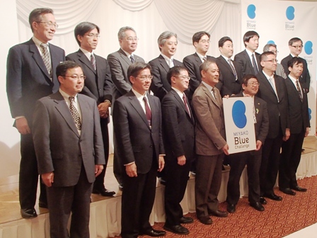 舞台上で関係者が2列に並び、そら豆のような形をした青色のデザインで「MIYAKO Blue challenge」と書かれたポスターを市長と関係者が手に持っている写真