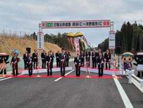 開通のお祝いの割れたくす玉を背景に、道路に敷かれたレッドカーペットの上で市長と関係者の方々計6人でテープカットを行い、それを多くの方々が見ている写真