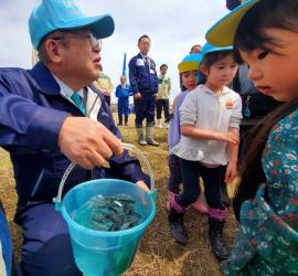 市長がたくさんの稚魚が入ったバケツを持ち、青い帽子を被った園児たちがそれを覗き込んでいる様子の写真