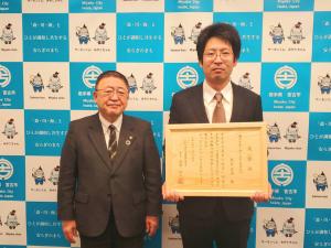 バックパネルの前で「いわて農林水産振興協議会長表彰・明日を拓く担い手賞」を受賞し額に入った賞状を両手で持っている田鎖憲幸さんと市長が並んでいる写真