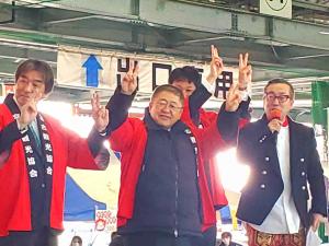 マイクを持った司会者の横で赤い法被を着た市長と魚市場関係者の方々が両手でピースサインをしている写真