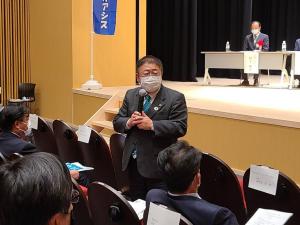 「みなとオアシスの集いin八幡浜」の前から二列目の自席から立ち上がった市長が、参加者の方を向いてマイクを持ち話をしている写真