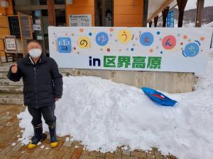 「雪のゆうえんち in区界高原」と書かれた看板と、その前に積もった雪の山の前で右手の親指を上に向けポーズをとる市長を写した写真