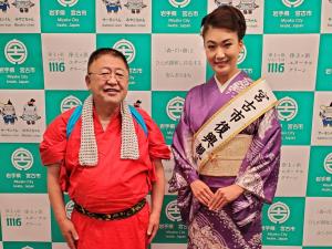 赤い着物を着て首にハチマキを掛けている市長と宮古市復興親善大使の市川由紀乃さんがバックパネルの前で並んでいる記念写真