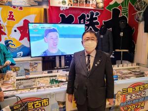 大漁旗が飾られた壁やモニターが置かれた施設で、「三陸・宮古市まるごと観光物産展」という文字やイラストがラッピングされた冷蔵ショーケースの前に立つスーツを着た市長の写真
