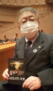 演奏会のパンフレットを両手で持ち、体を斜め横に向けて座って写真に写る市長の写真