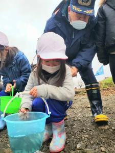 閉伊川の河川敷で、放流する前のさけ稚魚が入った水色のバケツを持った女児の後ろに立ち話しかけている市長の写真
