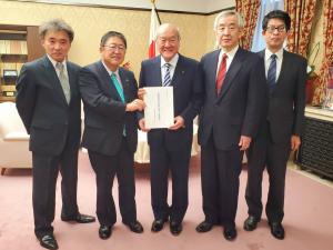 国旗や白い応接セットが設置された室内で、財務大臣の鈴木俊一氏と市長が一緒に陳情書を持って中央に立ち、3名の関係者が横一列に並んでいる写真