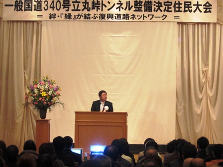 「一般国道340号立丸峠トンネル整備決定住民大会」の吊り看板が掲げられ、演壇に立つ市長と話を聞く参加者の写真