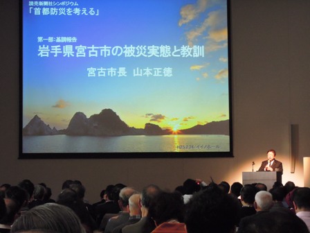 会場のホールに「岩手県宮古市の被災実態と教訓」と題した資料がスクリーンに資料が映し出されており、横の演壇に立ち、講演する市長と、講演に耳を傾ける参加者の写真