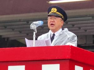 雨が降る中消防団員の制服と帽子を被り、その上にかっぱを着た市長が出初式での挨拶をしている写真